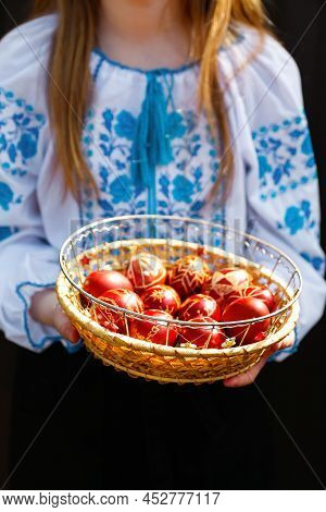 Defocus Ukrainian Girl In Vyshyvanka Holding Basket Of Colored Red Eggs On Black Background. Easter,
