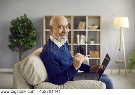 Happy, Smiling Senior Man Sitting On Couch At Home And Using His Laptop Computer
