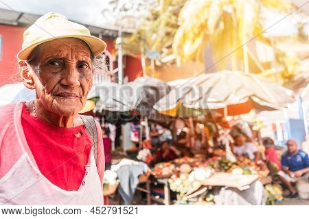 Poor Elderly Latin Woman Wearing Hat And Worn Clothes Shopping In A Traditional Market In Boaco