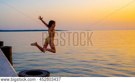 Girl Jumping From Pier In The Water At Sunset. Child Jumping In The Sea In Silba, Croatia.