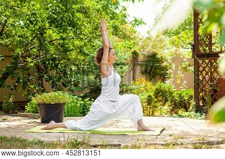 Young Woman Practices Yoga In The Summer Garden - Warrior Pose, Virabhadrasana I.