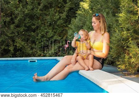 Summer Heat. Young Mother Holding Her Lettle Daughter. Baby Girl Drinks Water From A Bottle. Family 