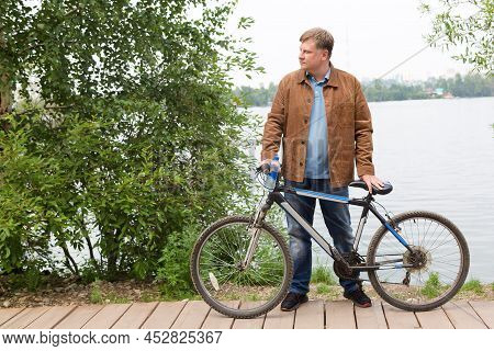 An Adult Blond Man In A Brown Corduroy Jacket Is Standing By The River Park With A Bicycle.