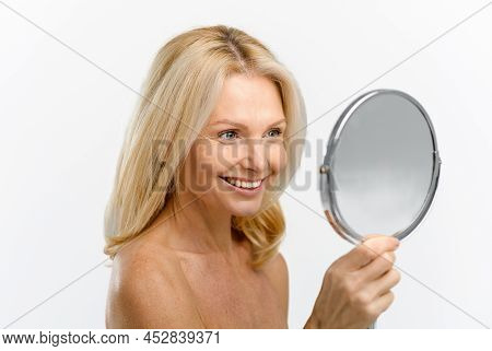 Cheerful Blonde 50s Woman Examining Her Reflection In Mirror Isolated On White. Middle-aged Female A