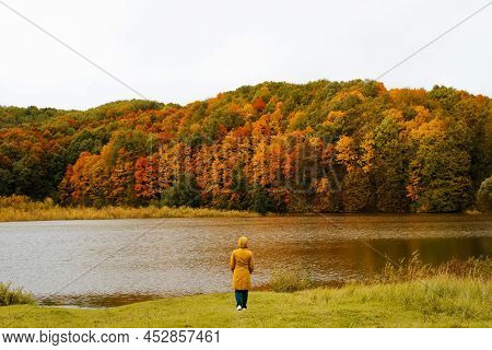 Back View Of Lonely Woman In Yellow Hooded Raincoat Standing By Lake Enjoying Autumn Scenery Outdoor