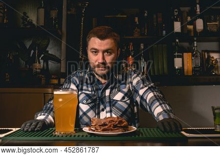 Bartender Standing Behind Bar Counter In Pub With Glass Of Beer And Plate Of Snacks
