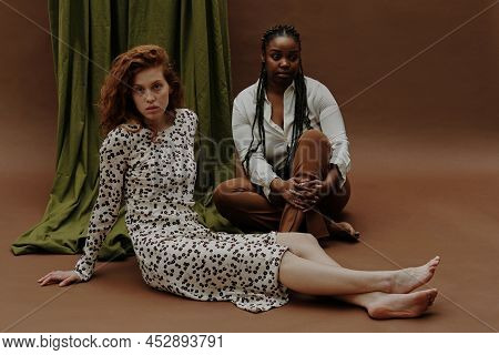 Portrait Of Two Diverse Young Women Sitting And Looking At Camera Against Brown Background