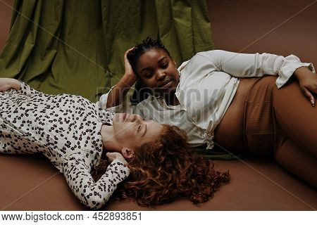 Portrait Of Two Women Of Different Types Relaxing On Floor Against The Green And Brown Background