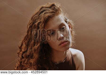 Young Woman With Curly Red Hair Looking Away And Thinking About Something Isolated On Brown Backgrou