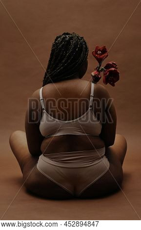 Rear View Of African Overweight Woman Sitting In Underwear And Holding Flower In Front Of Her Isolat