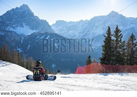 Rear View Of Tired Snowboarder Sitting On Snowy Landscape Against Mountains