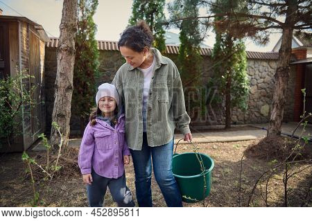 Young Pleasant Woman Holding A Bucket With Water, Standing With Her Cute Daughter And Watering Baby 