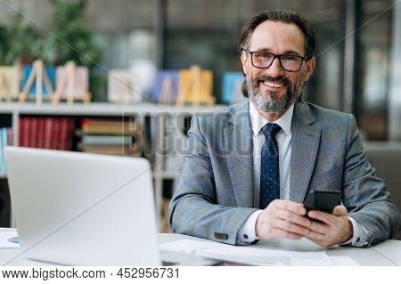 Smiling Grey-haired Business Man Looks Directly At The Camera, Sits At The Work Desk. Happy Influent