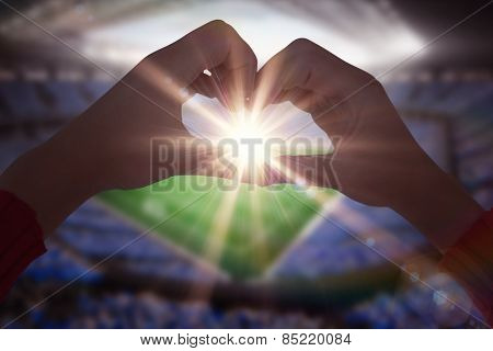 Woman making heart shape with hands against large football stadium with fans in blue