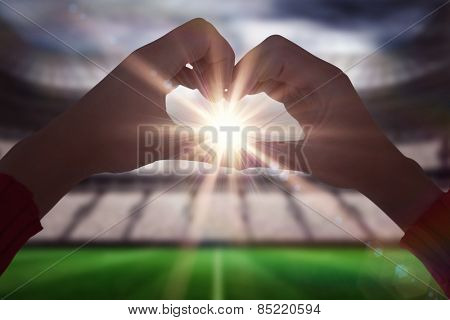 Woman making heart shape with hands against football stadium with fans in white