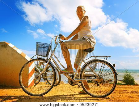 Young Woman Riding Bicycle Across River Bridge Next To Tropical Park