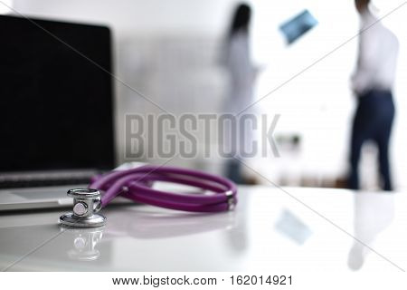 Laptop and stethoscope on the desk doctors standing in the background.