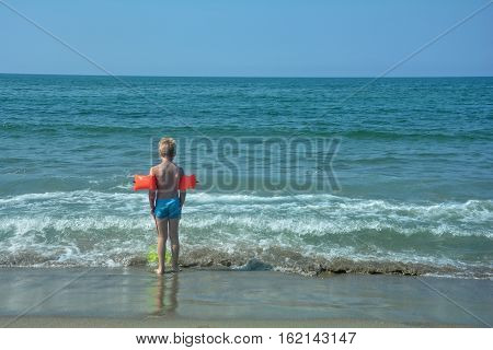Small blond boy with red swimming sleeves, stands on the beach and looks to the sea