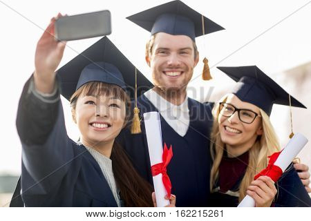 education, graduation, technology and people concept - group of happy international students in mortar boards and bachelor gowns with diplomas taking selfie by smartphone outdoors