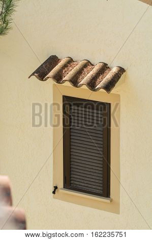 Window with brown closed shutters. Rain protection from roof tiles in a yellow painted house wall.