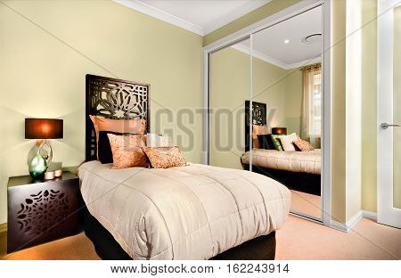 Single bed and the small table are made of wooden with carve. There is a table lamp on a cupboard and some glossy look fancy items on it. A huge mattress and few pillows on it. The mirror attached to green wall.
