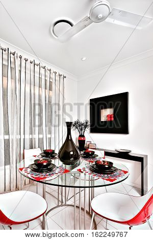 Dining room of a luxurious house chairs are curved and in red. The round table is made of glass and there is a shiny black vase with mugs and dishes on it a small black table can be seen behind the dining area