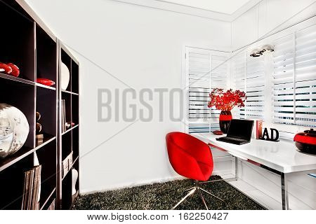 Work room walls are white and floor has a wool carpet. The chair is red. A wooden shelf with items the table is white near to the window. A laptop and shiny vases with flowers can be seen on the table with other ornamental items