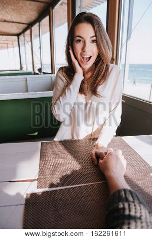 Vertical image of surprised woman sitting by the table on date in cafe near sea with hand on cheek and holding hand her man. First-person view