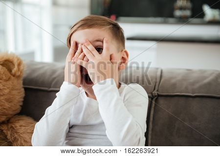 Picture of scared little boy on sofa with teddy bear at home watching TV while covering eyes.