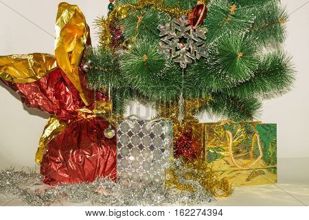 Christmas tree decorated with bright colorful toys snowflakes. Under the tree are gifts of silver and gold gift bags and gift in a red-golden shiny packaging
