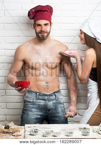 Pretty girl or beautiful woman cute helper sprinkles flour on handsome man or muscular chef cook baker with sexy muscle torso body with six packs and abs on kitchen wall