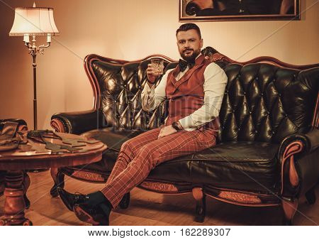 Extravagant stylish man with glass of whisky sitting on classic leather sofa in gentleman club
