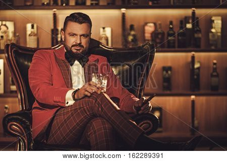 Extravagant stylish man with whisky glass sitting on armchair in gentleman club