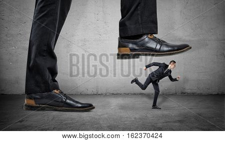 A businessman is running away from a big foot on a gray concrete background. Suppression of competitors. Big business.