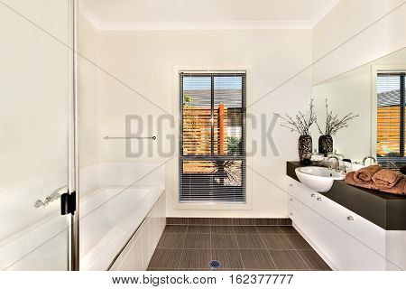 Luxurious bathroom of a modern house or hotel including white and long bath tub next to the countertop which is dark gray color and a white washstand with a silver faucet fixed to it front of the big and wide mirror.
