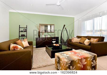 There is a puff box or cube chair made in textile. Dark color sofas and pillows next to a glass surface table made in wooden cover holding a fancy items in a dish. There is a wooden cupboard near the wall mirror. Modern kitchen in the luxurious house with