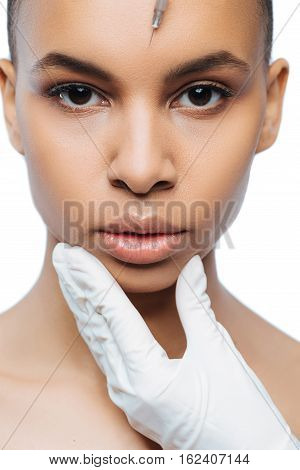 Victim of modern tendencies. Concentrated motionless young Negroid woman standing against white background and expressing concentration while receiving the injection in her forehead