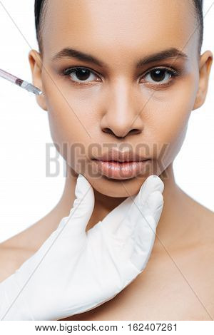 Beauty injection. Decisive motionless young Negroid woman standing against white background and expressing peacefulness while making the injection in her face