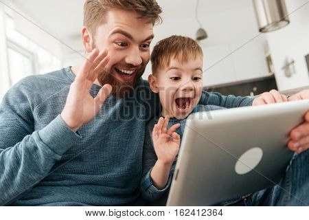 Image of cheerful father talking with friends by tablet computer and waving with his little cute son.