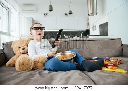 Image of little boy on sofa with teddy bear at home watching TV with 3d glasses while eating chips. Holding remote control.