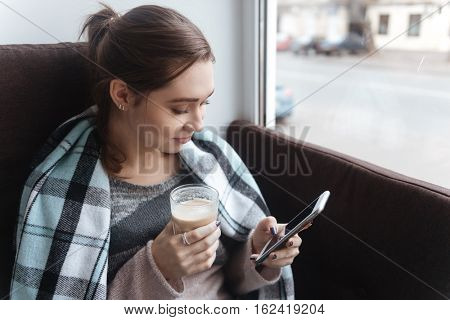 Photo of tenderless woman sitting in cafe near window under plaid while using phone. Look at phone.