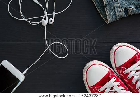 Smartphone with headphones and red sneakers on a black wooden surface top view