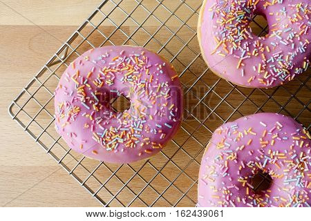 Closeup of three pink berry flavoured ring doughnuts with multi colored sprinkles. Positioned on stainless steel cooling rack with wooden background. Focus on doughnut to the left.