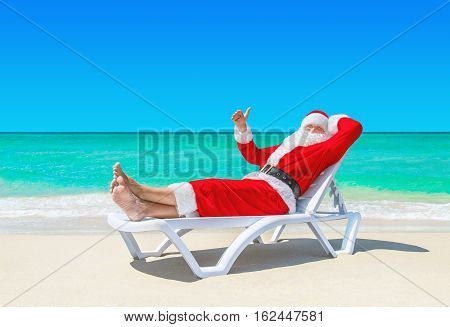 Santa Claus relaxing thumbs up gesturing on sunlounger at ocean tropical sandy beach against turquoise waves. Merry Christmas and Happy New Year travel destinations for tropical vacations concept.