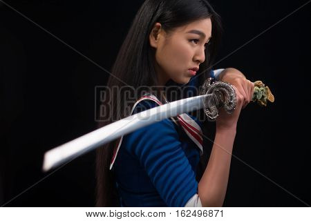 Pretty brunette lady with red lips looking like warrior. Asian lady posing sword for photographer in studio.