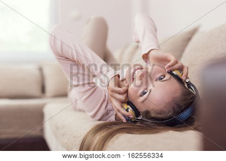 Young attractive woman lying on a couch in the living room wearing a headset listening to the music and enjoying her freedom and leisure time