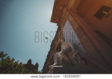 Marble statue of sitting man near facade of old historical building in park of Barcelona with flying pigeon behind sunny summer day teal sky Spain