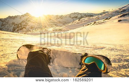 Snowboarder sitting at sunset in generic mountain ski resort - Winter sport concept with person on top of the mountain ready to ride down - Legs view point with warm backlighting filter