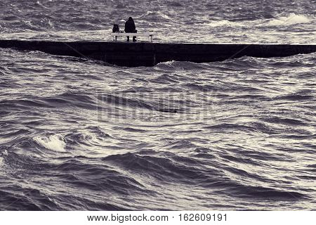 Lone fisherman on pier. Man sitting on deck chair in middle of stormy sea. Beautiful surging waves on water. Fishing in turbulent weather. Loneliness and solitude. Toned black and white color processing. Nice soft warm light