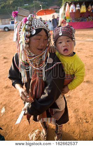 Mae Salong, Thailand - January 28, 2008 : 
In the mountains of northern Thailand. An woman from the Akha ethnic group carries her baby behind her back. To save her money Akha women sew silver coins on headdress.